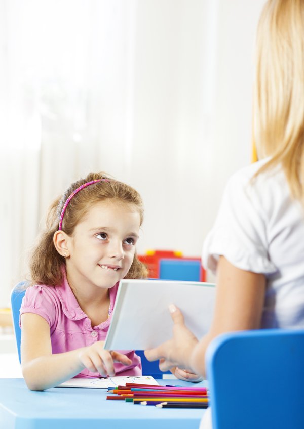 Cute little girl having speech therapy with young female speech/language therapist in a office. Therapist showing illustration of different objects to hear how she pronouncing some words. Selective focus to cute little girl looking at therapist and smiling.